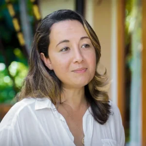 A woman in a white shirt with shoulder-length hair standing outdoors, looking reflective