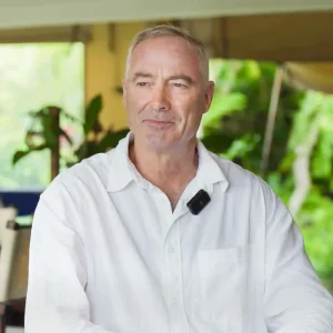 A man in a white shirt wearing a microphone, sitting in a relaxed indoor setting with greenery in the background
