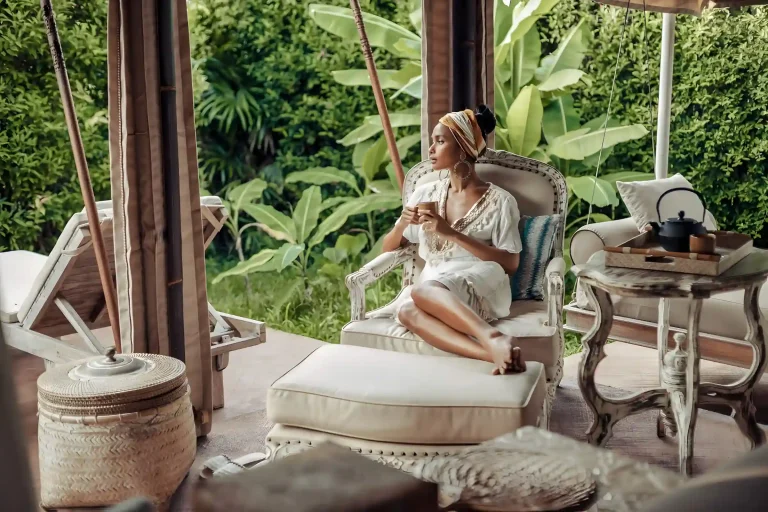 A woman in a light outfit relaxing on a patio chair, surrounded by lush greenery and stylish decor