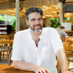 A man in a white shirt sitting at an outdoor dining area, smiling thoughtfully