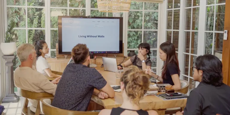 A team of professionals in a glass-walled meeting room discussing a project, with 'Living Without Walls' displayed on a large screen.