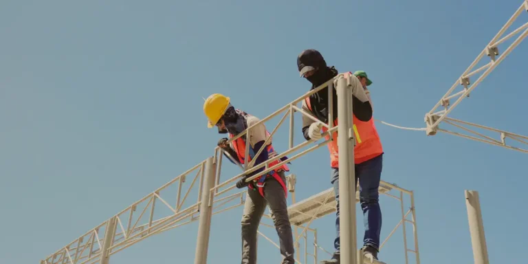 Two construction workers in safety gear assembling a tent structure under a clear blue sky