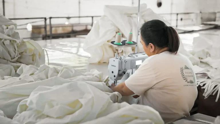 A seamstress working on a large piece of white fabric using a sewing machine in a workshop setting